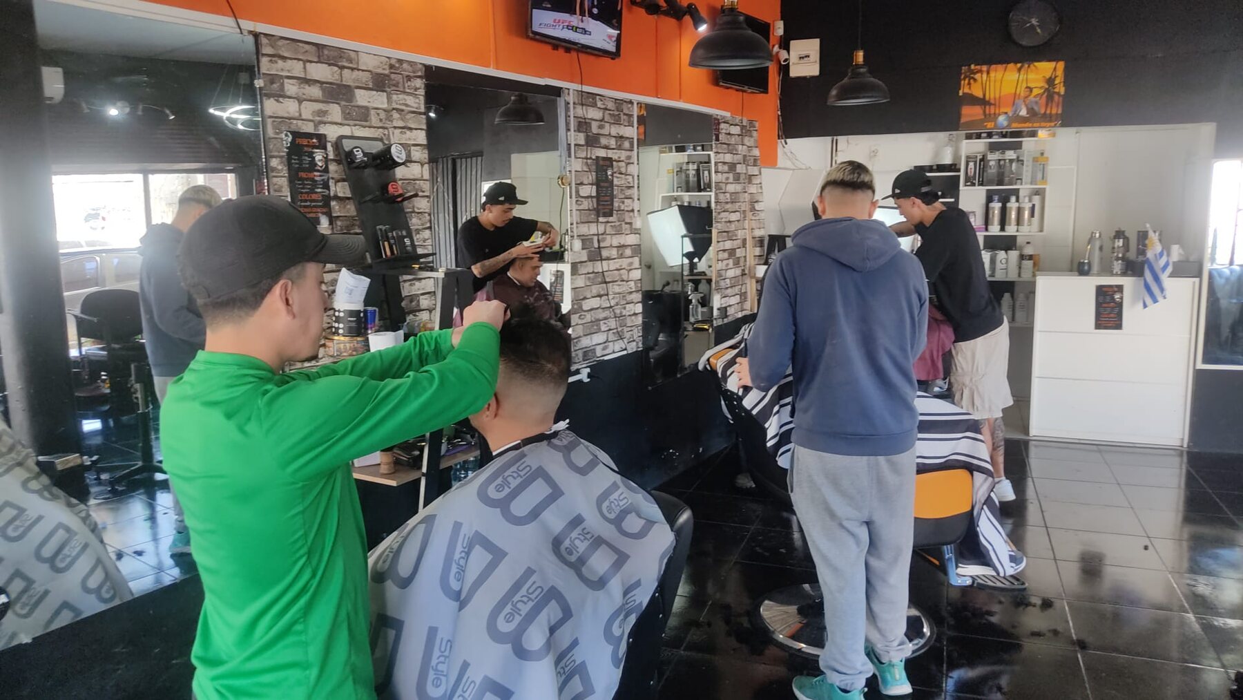 El salón lleno un mediodía: tres barberos trabajando, los espejos, la bandera de Uruguay sobre la cómoda blanca y el televisor prendido.
