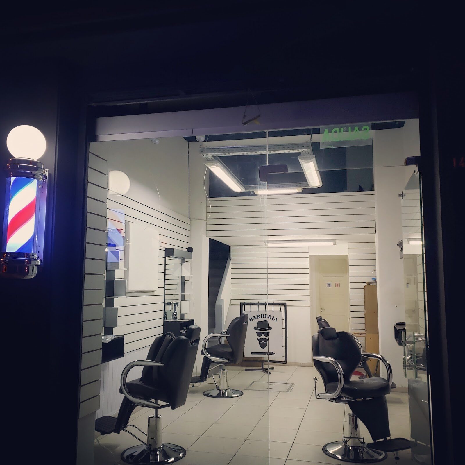 El frente del local de noche: paredes de listones blancos, tres sillones negros y el poste de barbero prendido junto a la puerta.
