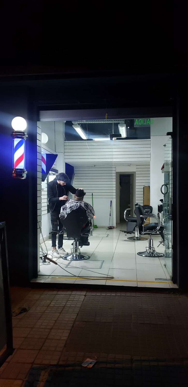 La barbería de noche, vista desde la vereda de Yatay: el poste de barbero prendido en la puerta y adentro, bajo los tubos, un barbero cortando.