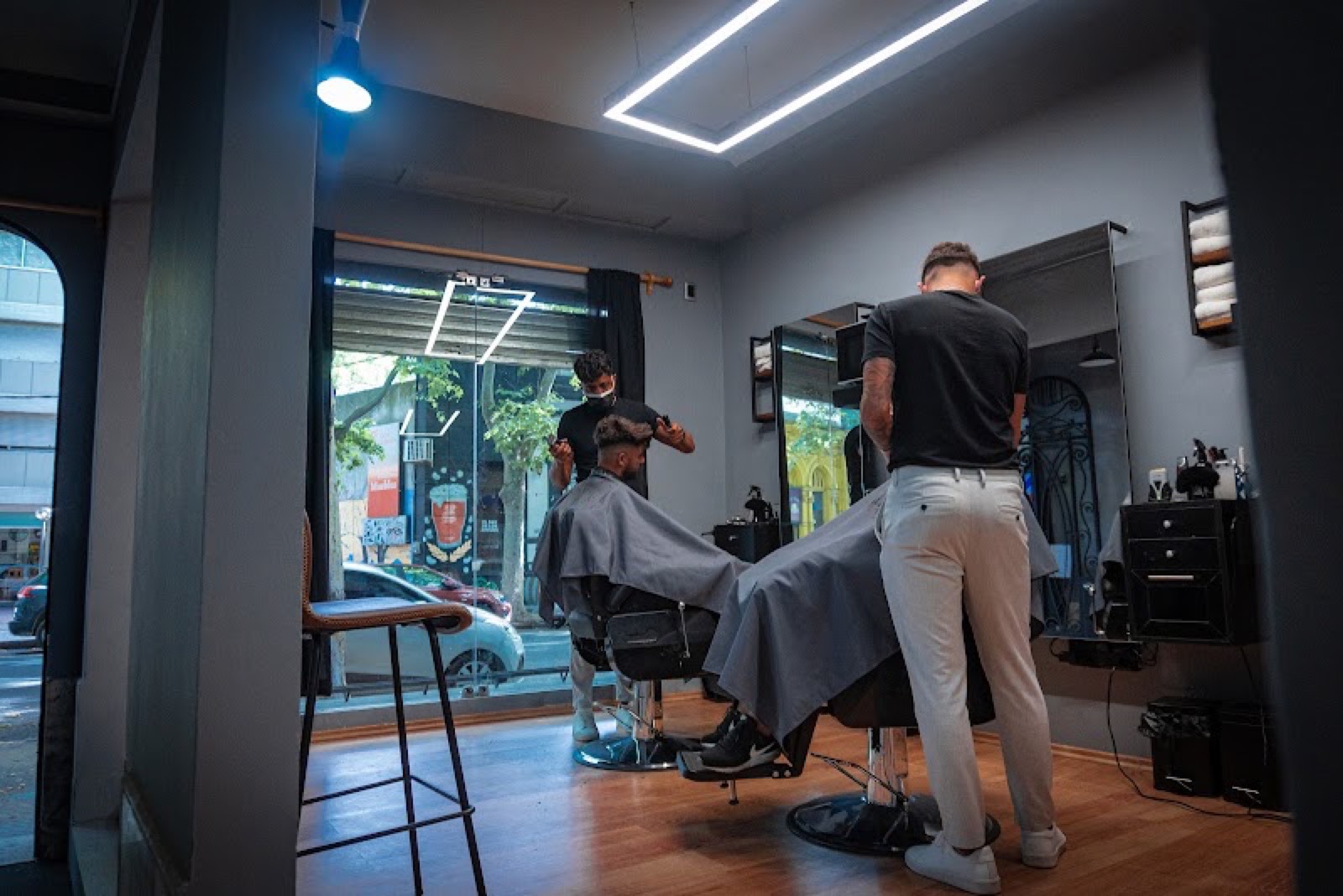 Dos barberos trabajando en el salón de Maldito, con la vidriera a la calle al fondo.
