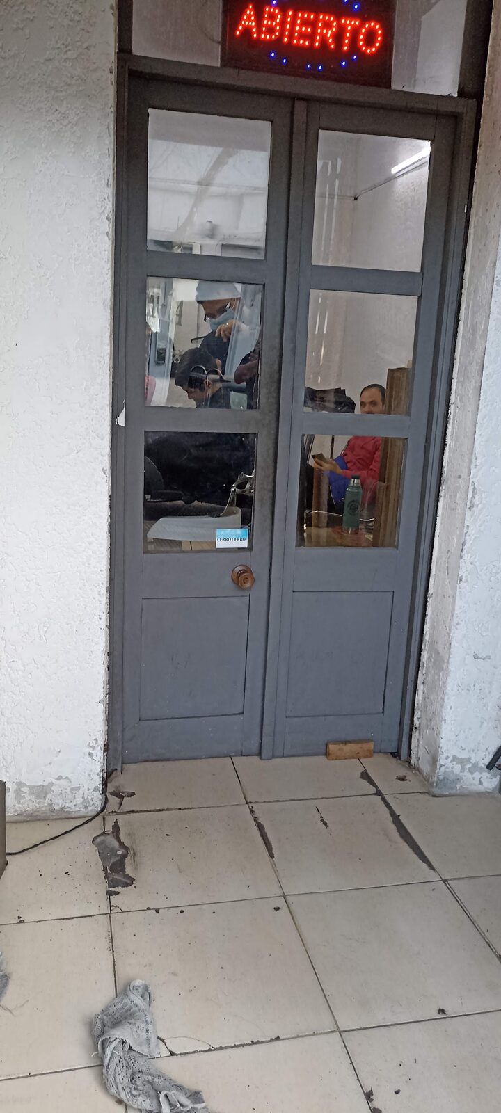 Desde la vereda se ve, a través del vidrio, a Leonel cortando y a un cliente esperando sentado.